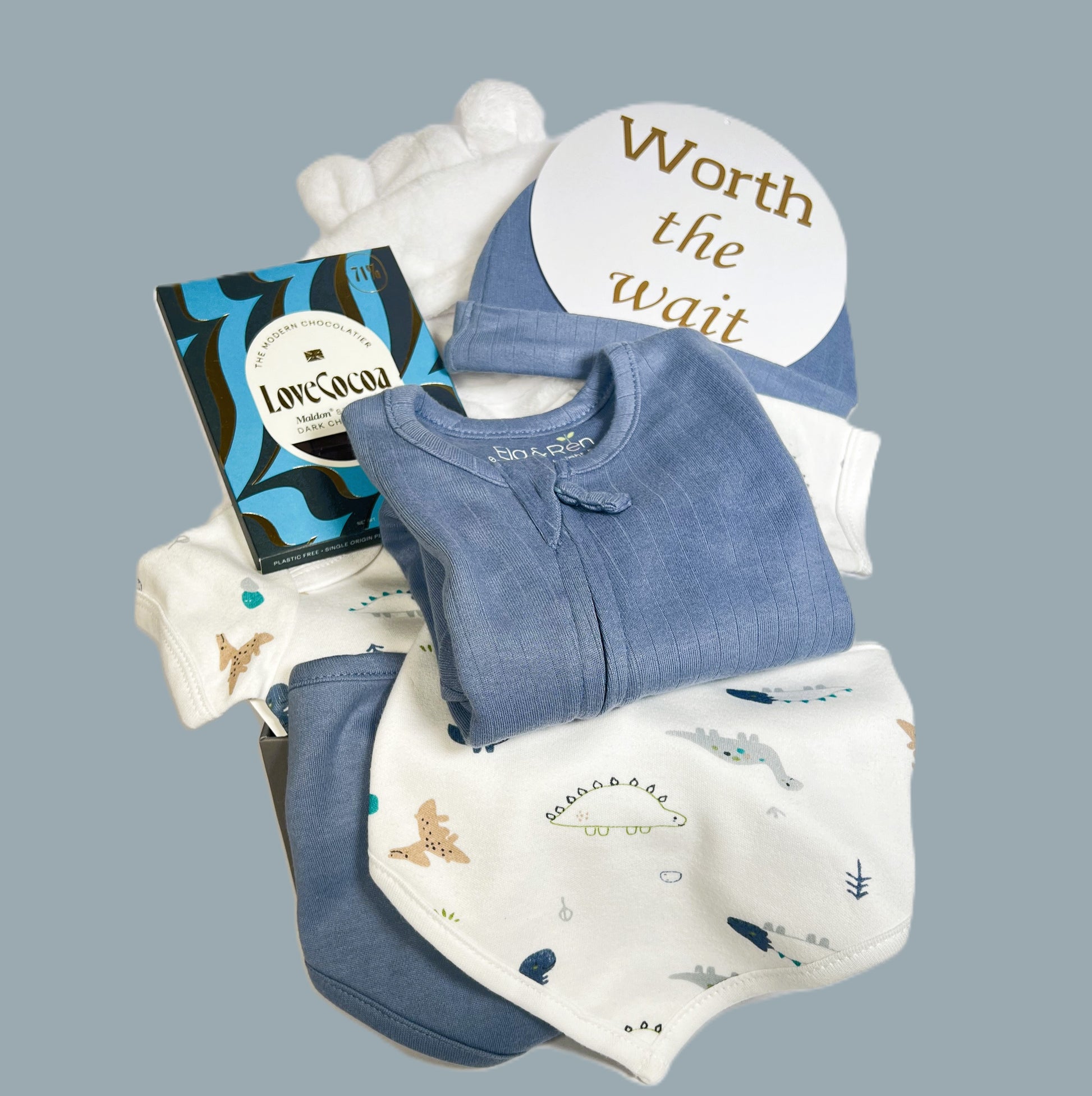 This baby boy hamper comprises a blue baby sleepsuit in organic cotton with a dinosaur print baby bodysuit and matching babby bib and a baby hat, a white baby dressing gown, a bar of Love Cocoa chocolate and a baby keepsake box