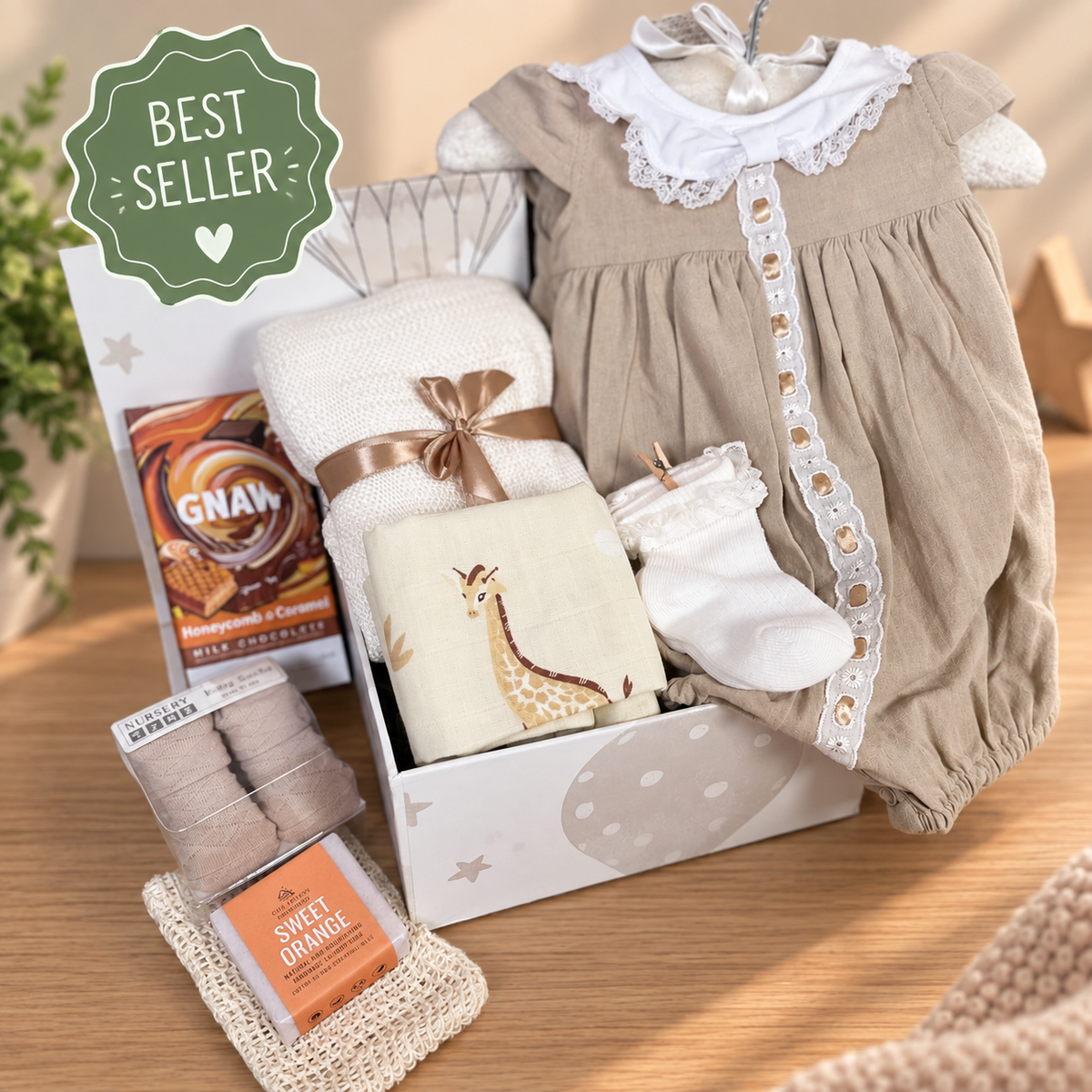 A baby gift hamper arranged on a beige background, including a beige and white baby outfit, a folded white bamboo baby blanket, giraffe-print muslin square, baby socks, and new mummy treats. 