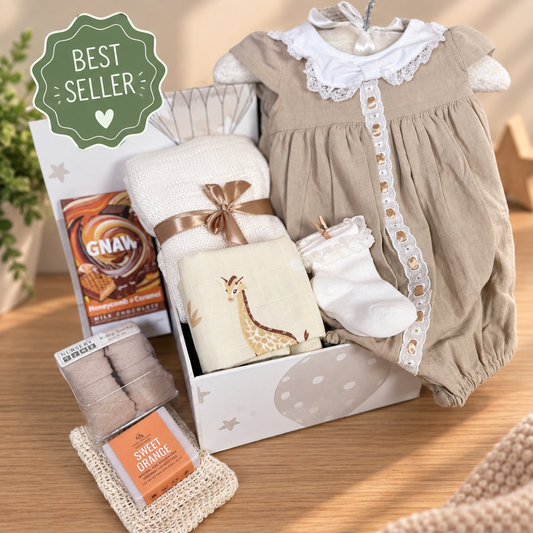 A baby gift hamper arranged on a beige background, including a beige and white baby outfit, a folded white bamboo baby blanket, giraffe-print muslin square, baby socks, and new mummy treats. 