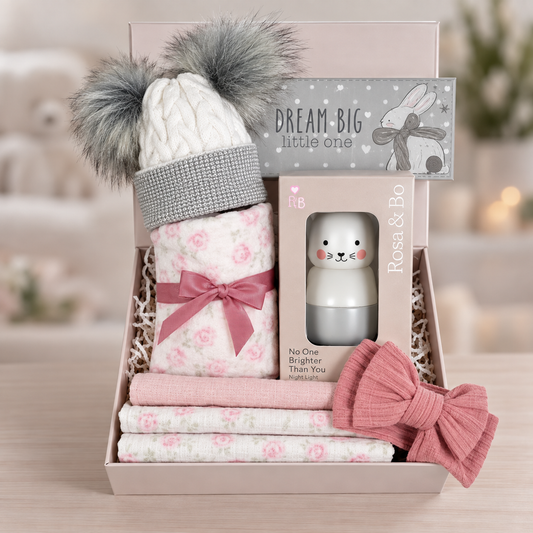 New baby gifts hamper with a pink rose bud baby blanket with cotton waffle to reverse and three matching baby muslins. A Rosa and Bo bunny stacking toy, a pink baby headband, a nursery plaque and a grey and white baby pompom hat.