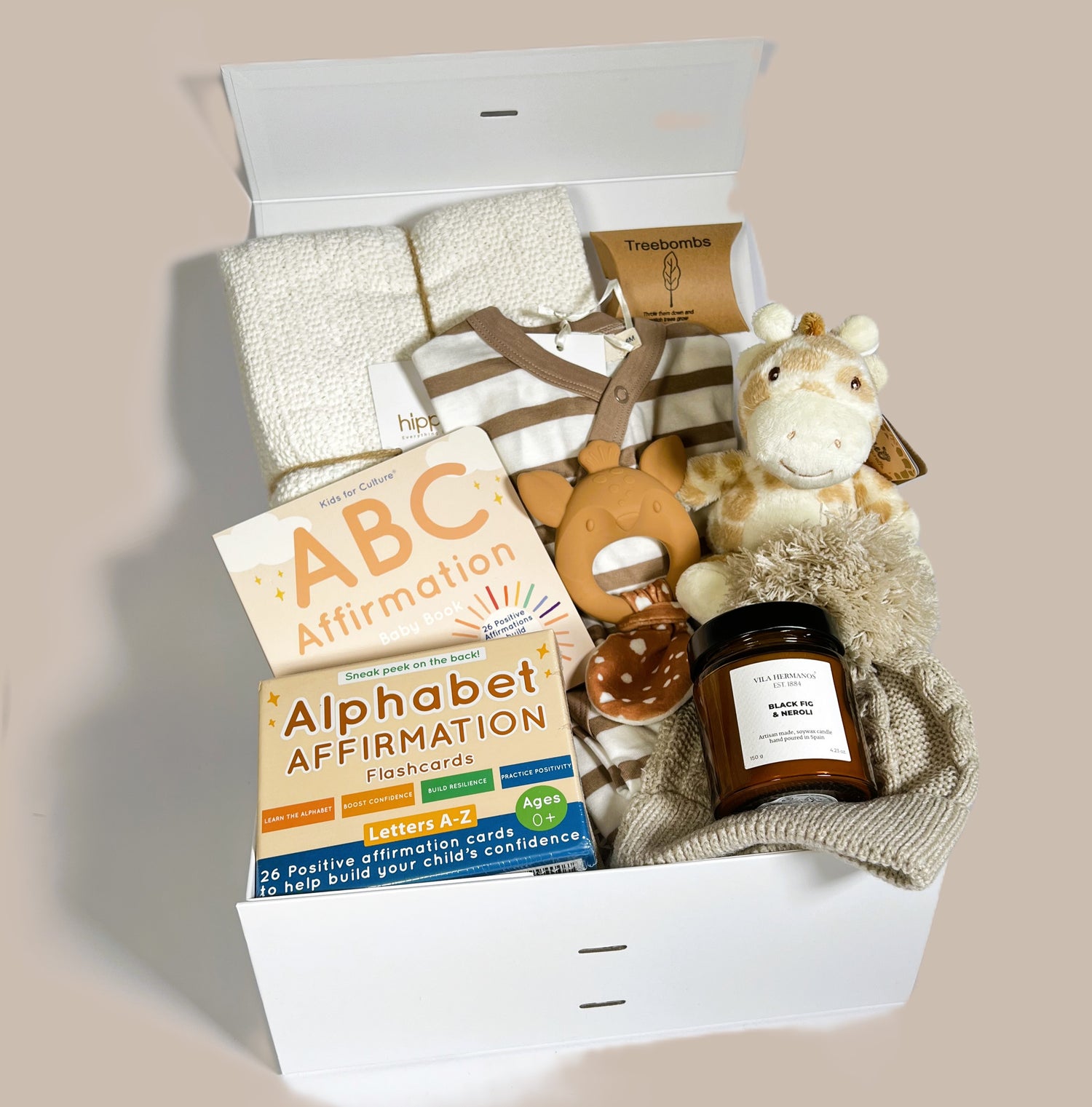 Baby hamper containing a white cotton cellular baby blanket. Organic cotton baby romper. A Ebba Gigi giraffe baby rattle, a baby pompom hat in taupe. A Leika baby teether a pack of affirmation flashcards and an baby board book. A fig and Neroli soya candle and 3 Treebombs.