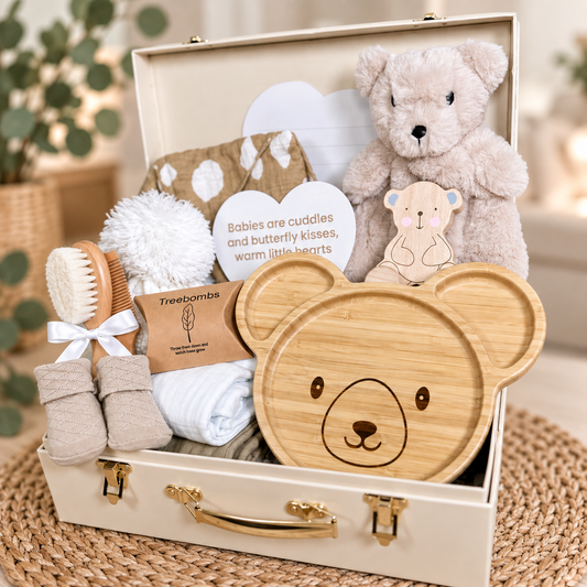 A curated baby gift hamper arranged inside an open cream-colored baby keepsake case. The set includes a plush teddy bear, a wooden bear-face tray, a wooden baby brush, a pair of beige booties, a folded white and taupe baby muslins, a brown patterned swaddle cloth, a heart-shaped nursery plaque, and a small wooden bear push along toy , a wooden baby hairbrush and comb set and a bear baby comforter .