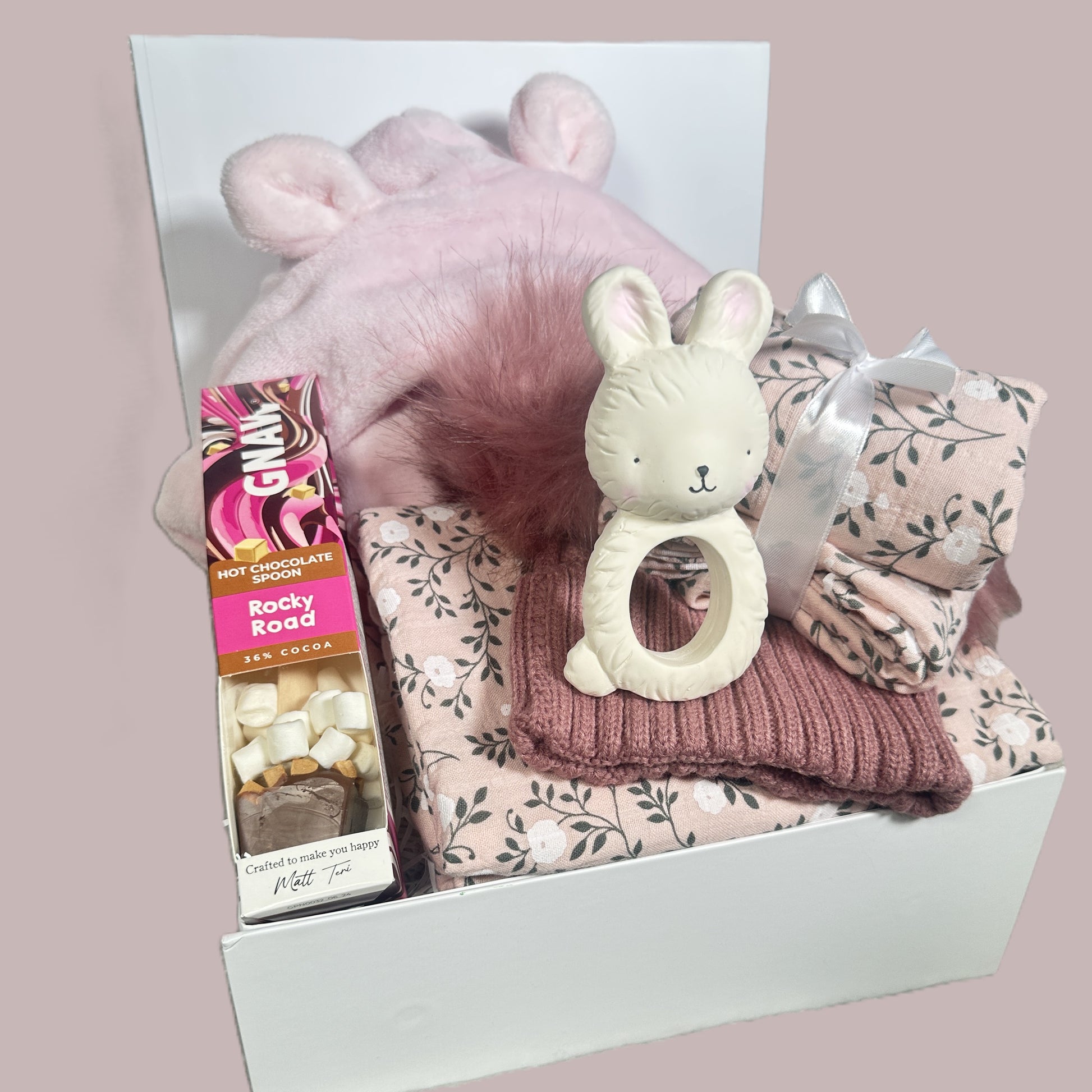 Gift box with baby items including a pink hat, teething ring, and blanket on a light pink background