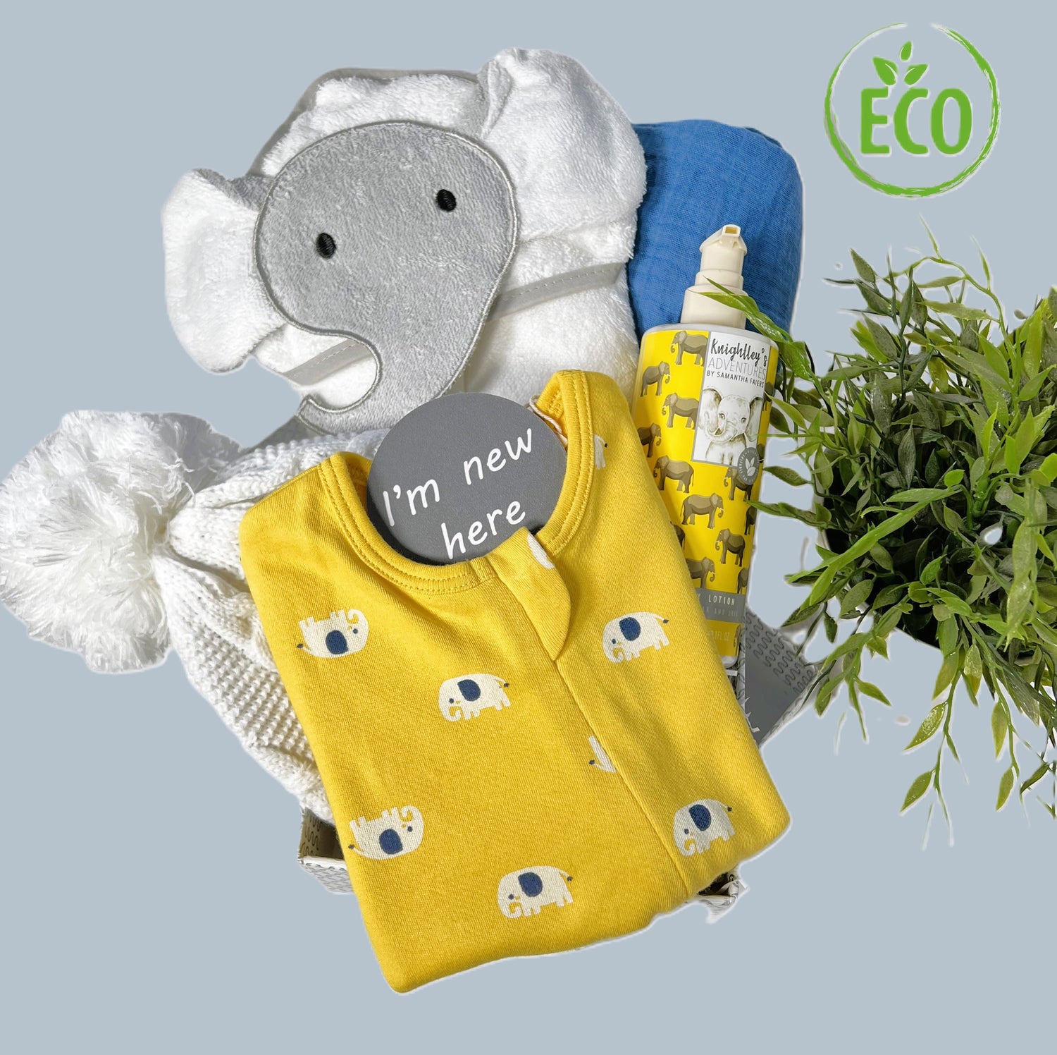 Elephant themed new baby gift with a white cotton hooded baby towel with grey elephant face, a organic cotton zipped baby sleepsuit in yellow with a navy blue and white elephant print, a bootle of paraben freen baby lotion, a very large baby muslin swaddle blanket , a white baby pompom hat all in a hand crafted silver and white baby keepsake box.