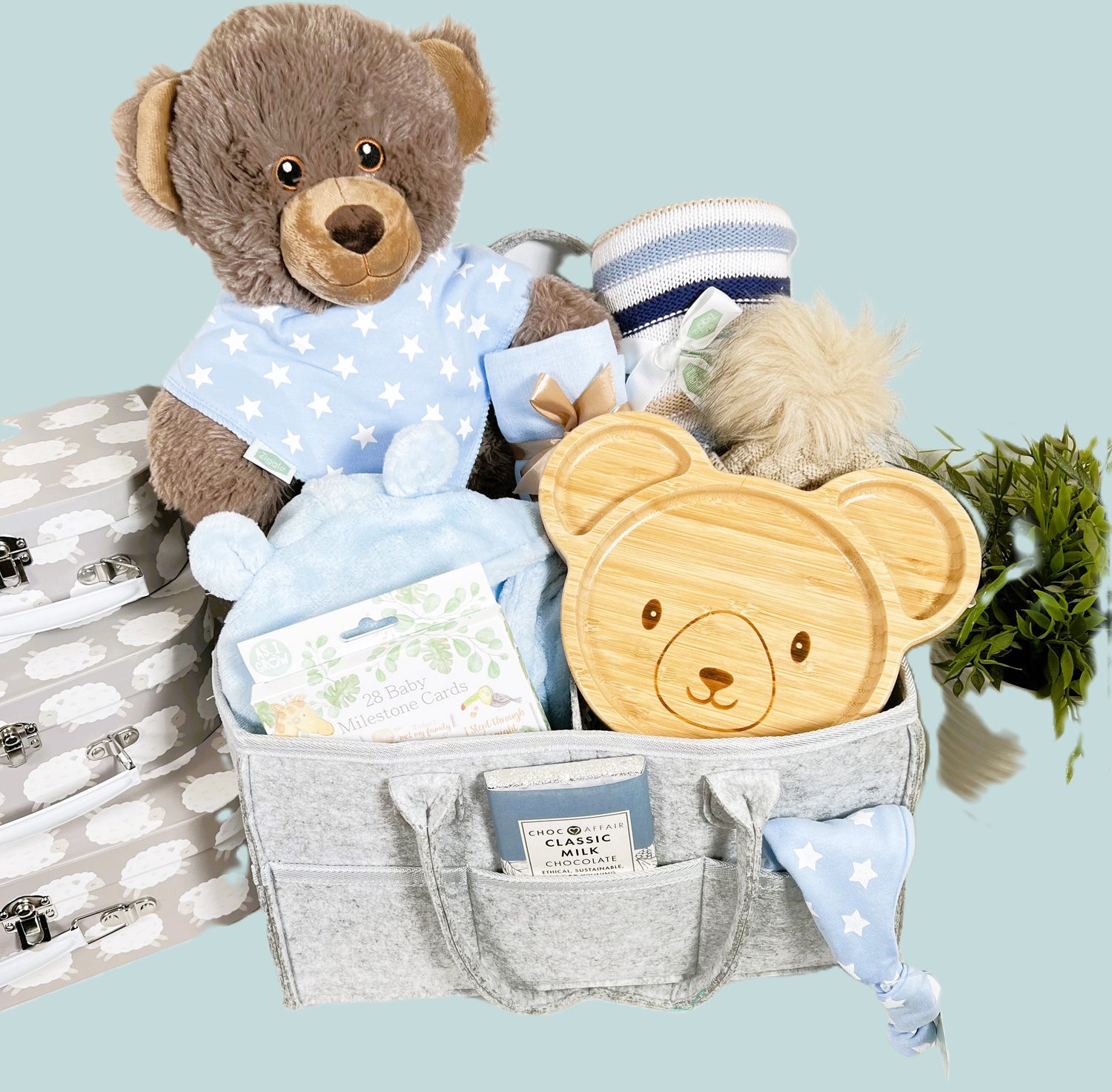 Nappy Caddy new baby boy gift in a fgrey felt nappy caddy with thre compartments and six pockets containing a large brown bear soft toy made from recycled plastics, a bamboo baby plate with bear face, a blue hooded baby dressing gown, a pack of baby milestone cards, baby pompom hat, a blue baby muslin square, baby toiletries and a Choc Affair bar of chocolate.