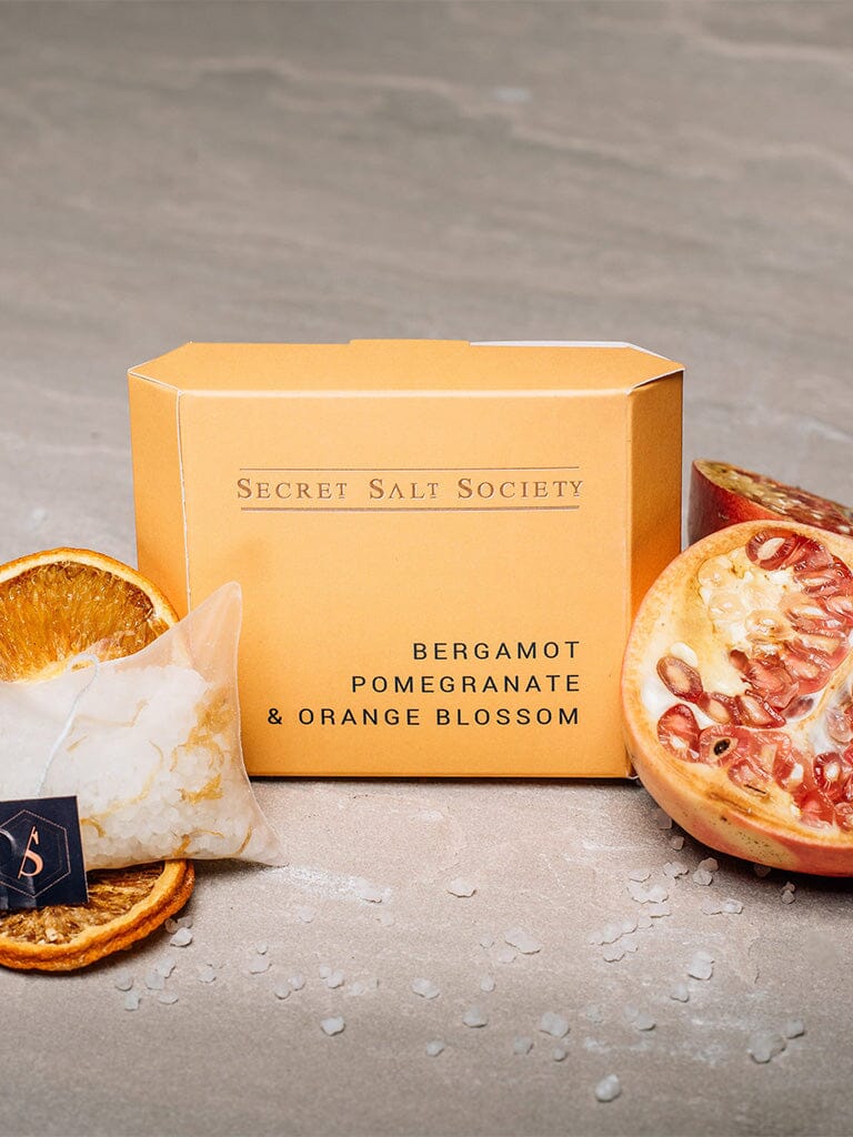 Box of Secret Salt Society Bergamot Pomegranate & Orange Blossom salt with salt crystals and pomegranate slices on a gray surface.
