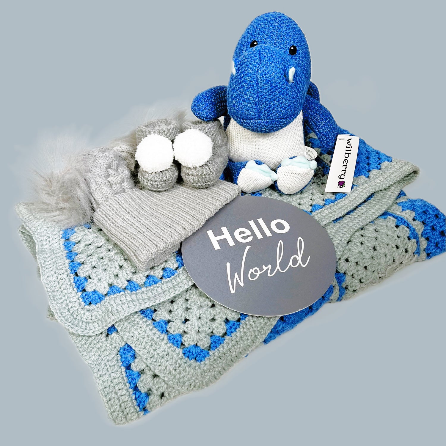Grey and blue granny square baby boy crocheted baby blanket with a pair of grey baby booties with pompoms, a grey baby pompom hat, a "Hello World" baby photography plaque.