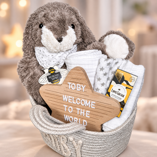 A baby gift basket set in a woven basket, featuring a grey plush bunny with a white bib, white muslin blankets, a wooden star sign reading 'TOBY WELCOME TO THE WORLD', and, set against a softly blurred warm indoor background