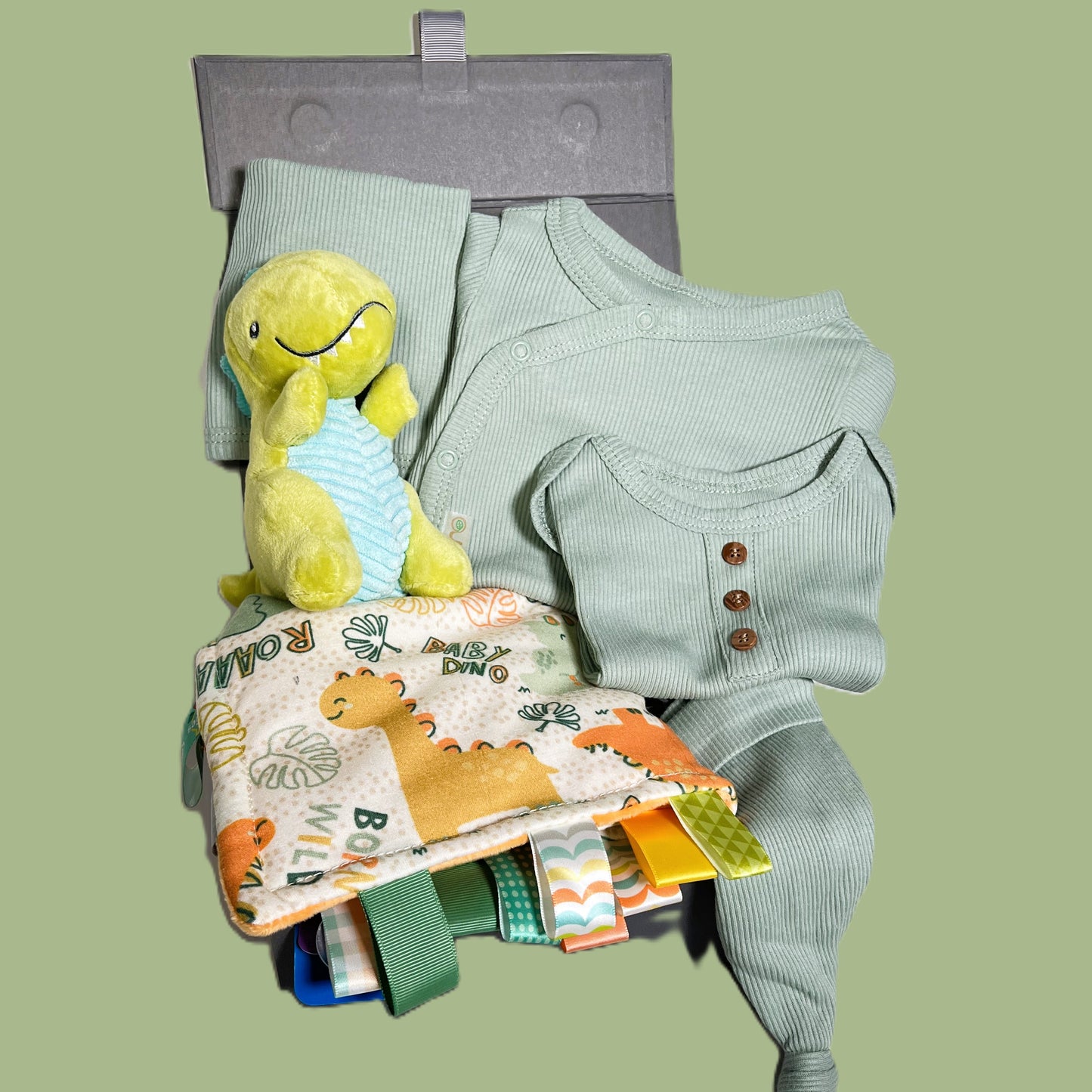 A dinosaur themed baby hamper gift with a Mary Meyer dinosaur print Taggie blanket, a Mary Meyer green dinosaur baby rattle and a sage green cotton layette set.