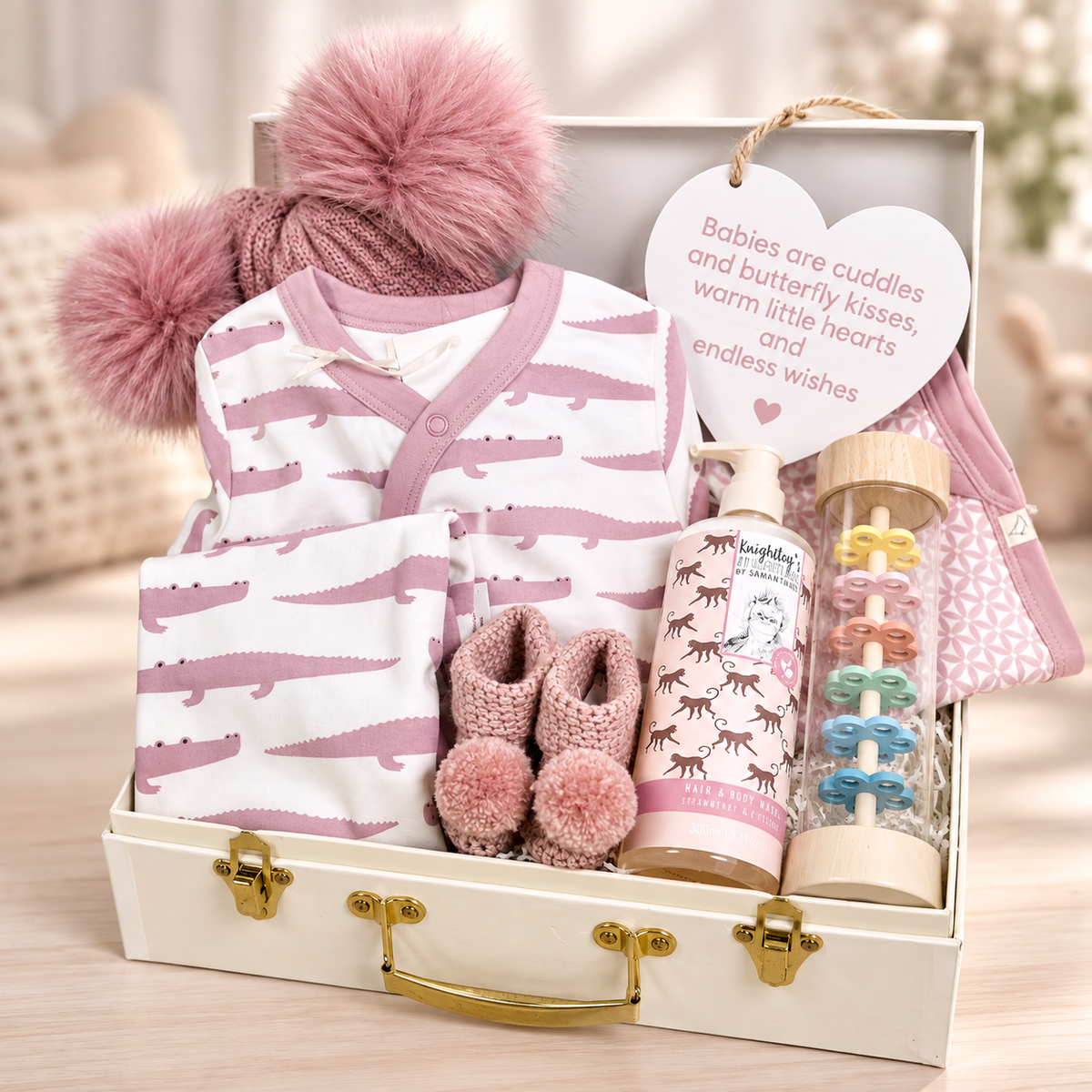 A  new baby gift gift arranged inside a cream baby keepsake case with gold-tone hardware. The set includes a pink and white crocodile-patterned baby outfit, a matching pink knit hat with a large pom-pom, folded pink and white baby bodysuit, a pair of pink crocheted booties with pom-poms, a bottle of baby bath, a colourful baby rainmaker toy, and a heart-shaped nursery plaque that reads, 'Babies are cuddles and butterfly kisses, warm little hearts and endless wishes.