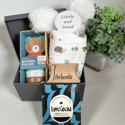 A Lovely new baby gift containing a eco friendly wooden bear stacker toy, a large muslin with jungle print, a white pompom baby hat, a bar of Love Cocoa chocolate, 3 Treebombs and a baby photography disc in a grey square baby keepsake box.