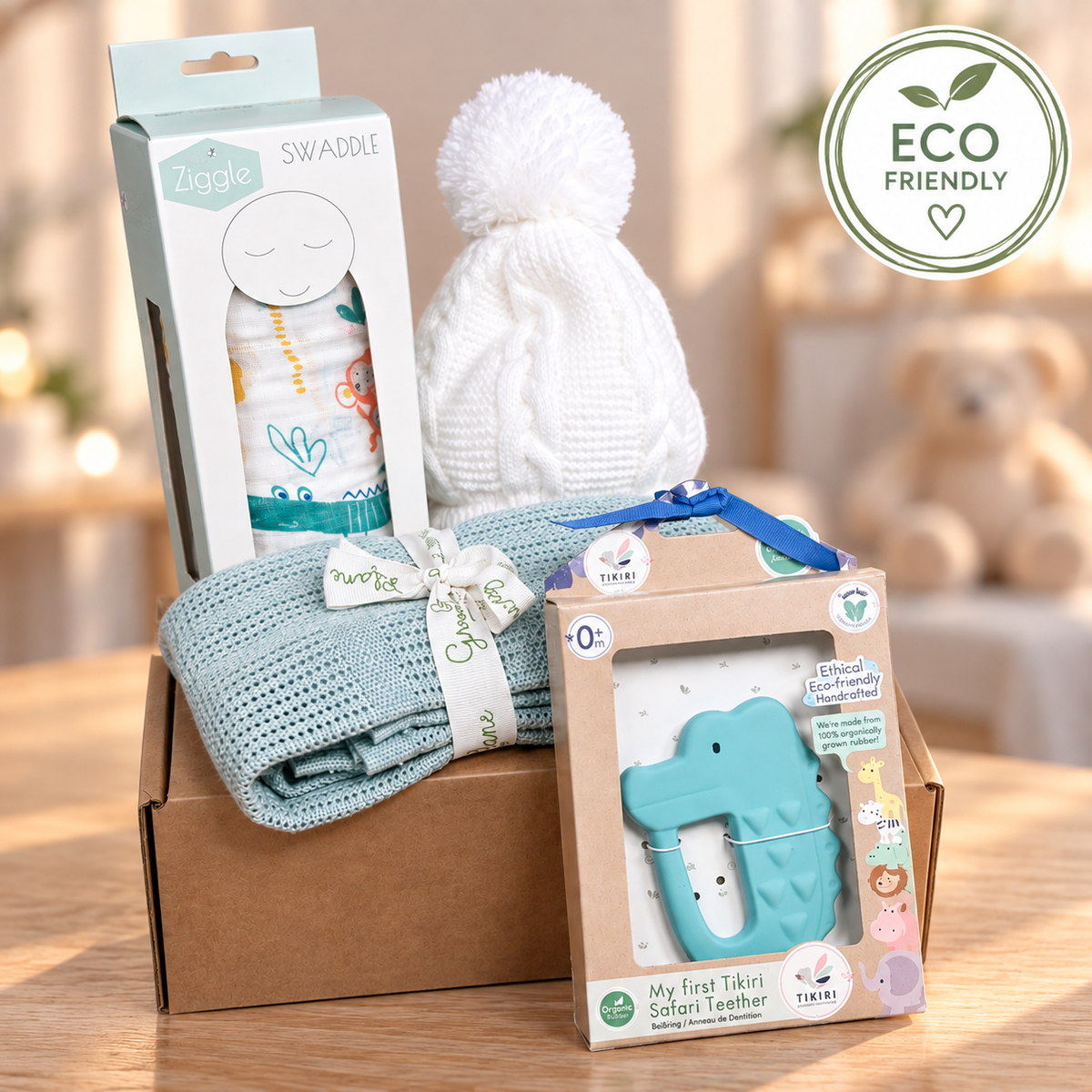 A baby gift set arranged in a cardboard box on a wooden surface, featuring a white knit pom-pom hat, a white swaddle blanket with a fun jungle print,  and a Tikiri 'My First Teether' safari natural rubber teether and a sage bamboo cellular baby blanket. An 'ECO FRIENDLY' badge is visible in the top right corner.