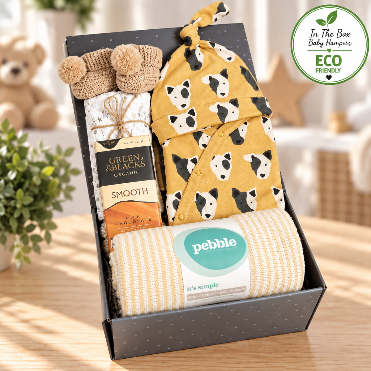 Mustard and cream unisex new baby gift hamper containing a GOTS certified organic cotton baby romper with a dog face print, fold over cuffs and feet with a matching baby knot hat, a hand knitted cotton baby blanket, an elephant print baby muslin, a pair of crocheted baby booties, a bar of green and Blacks organic "smooth" chocolate all in a grey magnetic baby keepsake box.