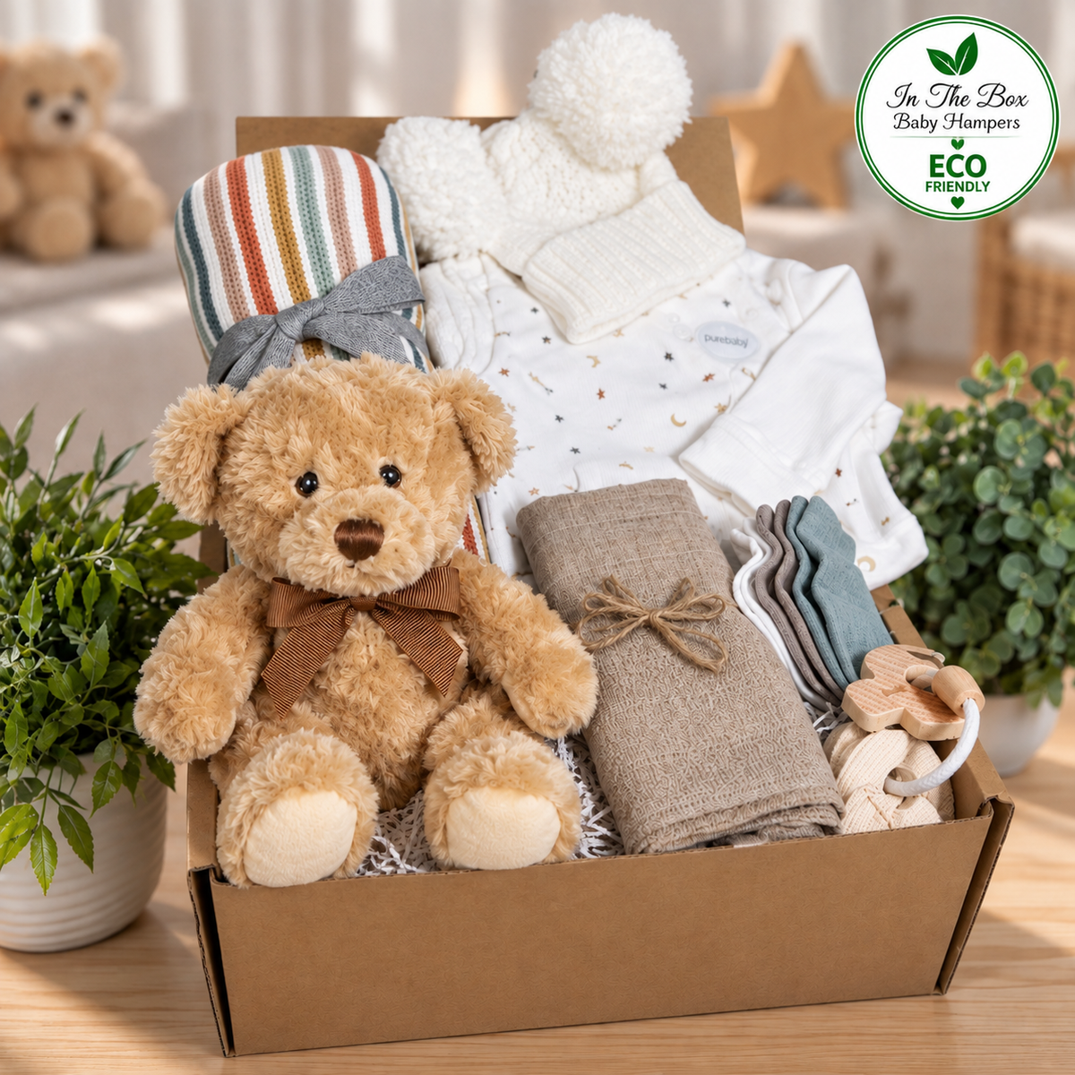 A baby gift box filled with baby essentials including a teddy bear, a striped baby blanket, a white knit hat with a pom-pom, a onesie, folded baby muslin, and a wooden teething toy, set on a wooden surface with potted plants and a brand badge in the top right corner.