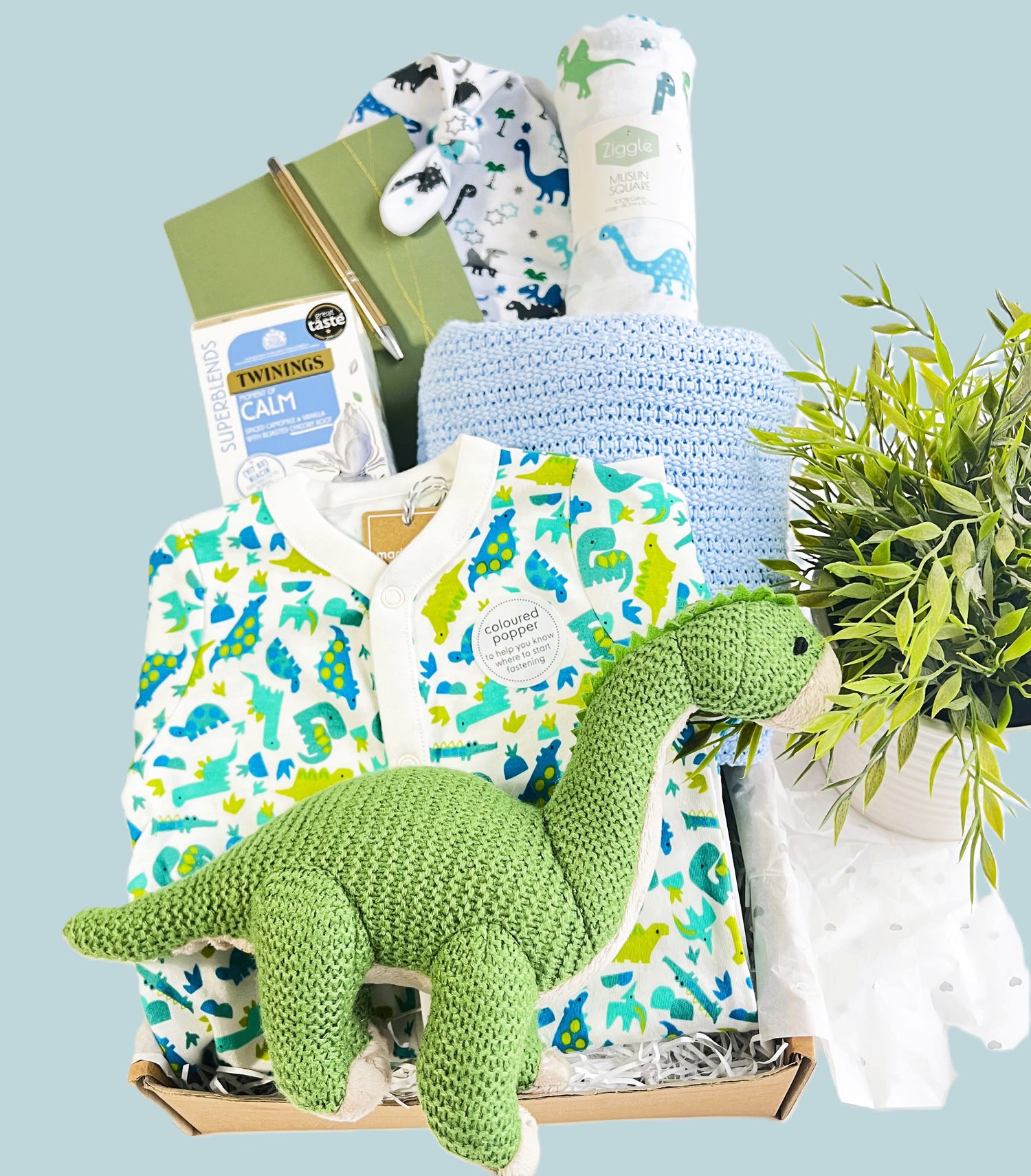 Newborn baby boy hamper gift, dinosaur themed with a green Wilberry medium brontosaurs baby toy, dinosaur print baby sleepsuit, blue soft cotton cellular baby blanket, calming tea for the new parents, a baby journal and baby knot hat and muslin square.