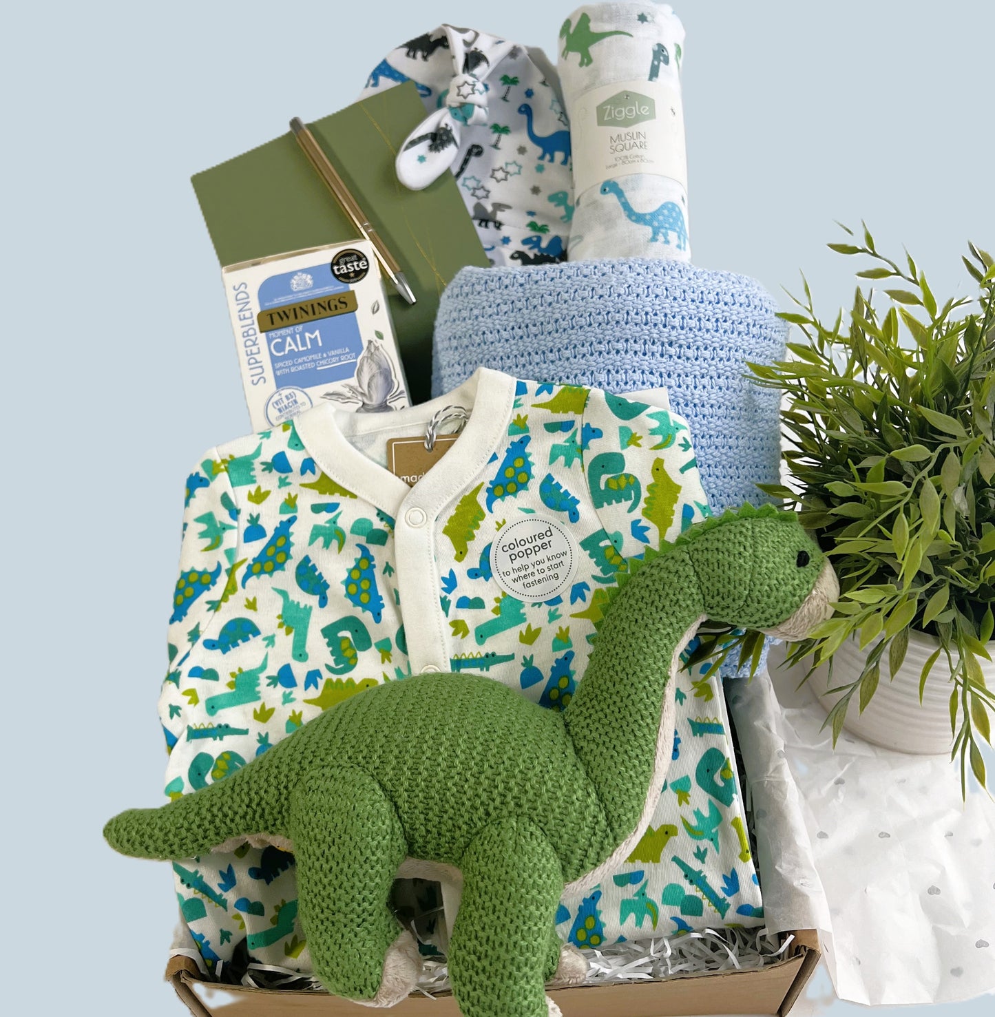 newborn baby boy hamper gift, dinosuar themed with a green Wilberry medium brontosaurs baby toy, dinosaur print baby sleepsuit, blue soft cotton cellular baby blanket, calming tea for the new parents, a baby journal and baby knot hat and muslin square.