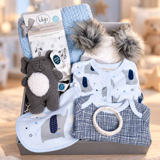 A curated baby boy gift hamper in a grey baby keepsake box featuring a light blue cotton cellular baby blanket, a matching elephant-print bodysuit, a navy-and-white check bodysuit with a silicone teething bangle, a plush grey elephant baby rattle, a bib, and a baby pom-pom hat. 