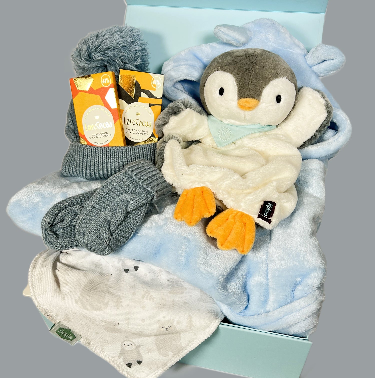 Baby boy hamper in a blue magnetic baby keepsake box containing a Kaloo penguin baby comforter, a grey baby pompom hat and mittens, 2 bars of Love Cocoa chocolate, a blue baby dressing gown an a cotton baby dribble bib.