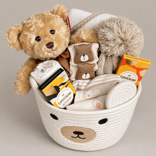 A teddy bear-themed gift basket filled with plush toys, blankets, and packaged treats. The basket is cream-colored with a bear face design, and it is filled with a large brown teddy bear, baby socks with bear faces , folded white and beige babywear, a pom-pom hat, and 2  chocolate bars and a hooded baby towel, set against a plain beige background.