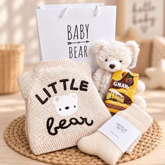 A baby gift set arranged on a woven mat, featuring a cream knit sweater with 'LITTLE bear' and a bear face, a white teddy bear, a white gift bag reading 'BABY BEAR', cream socks, and a GNAW chocolate bar.