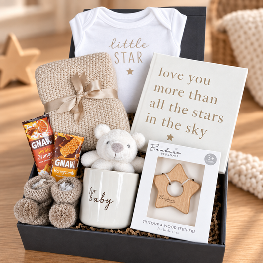A baby gift box set containing a white onesie with 'little star' text, a cream knit blanket tied with a ribbon, a card reading 'love you more than all the stars in the sky', GNAW orange and honeycomb cookies, a cream mug with 'baby' text, a plush teddy bear, and a package of Bumbo wooden teething toys, arranged in a black box on a wooden surface.