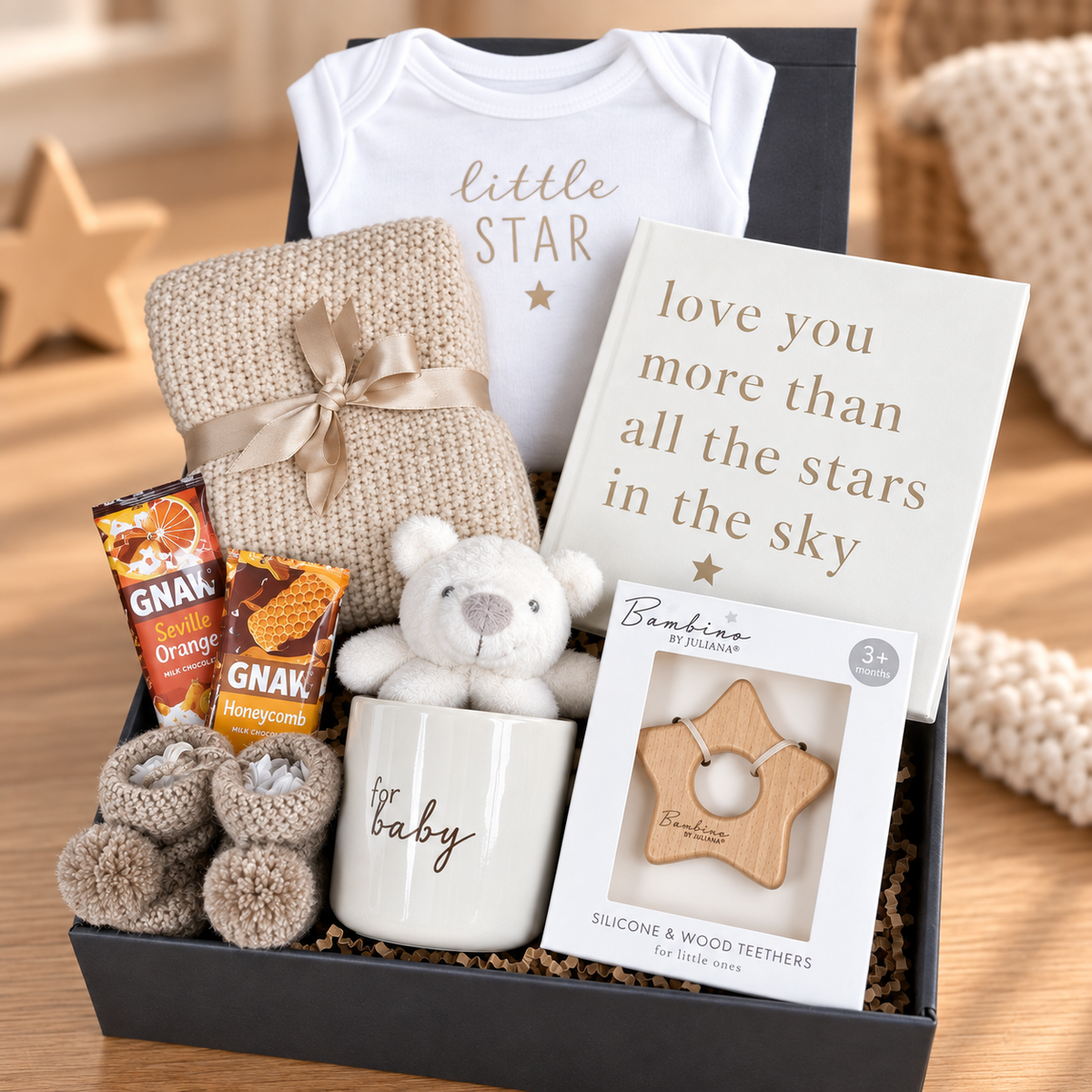 A baby gift box set containing a white onesie with 'little star' text, a cream knit blanket tied with a ribbon, a baby photo album reading 'love you more than all the stars in the sky', GNAW orange and honeycomb chocolate bars, a cream mug with 'baby' text, a plush teddy baby rattle, and a package of Bambino wooden teething toy, arranged in a grey baby keepsake box on a wooden surface.