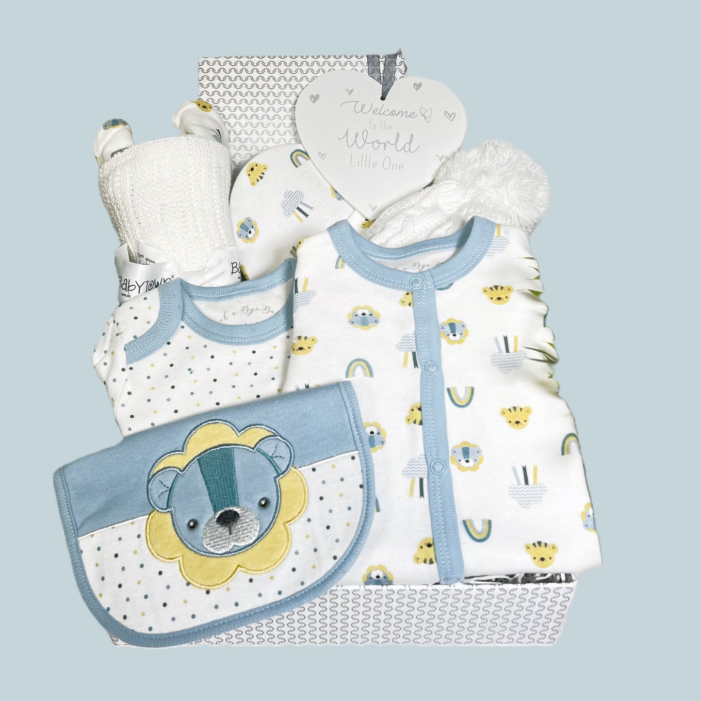 A new baby boy baby shower gift in a silver  and white baby keepsake box containing a cotton layette set with blue and yellow lion print, a white cotton cellular baby blanket, a baby pompom hat and a grey and white heart shaped nursery sign.