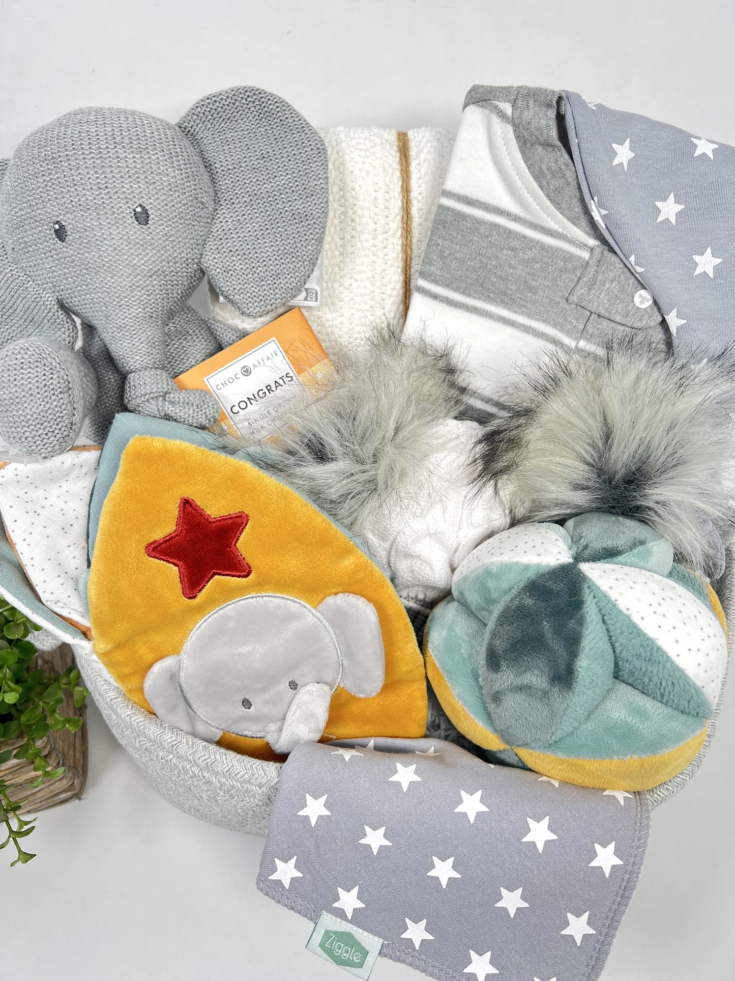 Luxury baby shower gift containing a knitted elephant toy, a cloth sensory book and matching activity ball, a cellular baby blanket, baby pompom hat, zipped baby sleepsuit and a baby bib and baby knot hat.