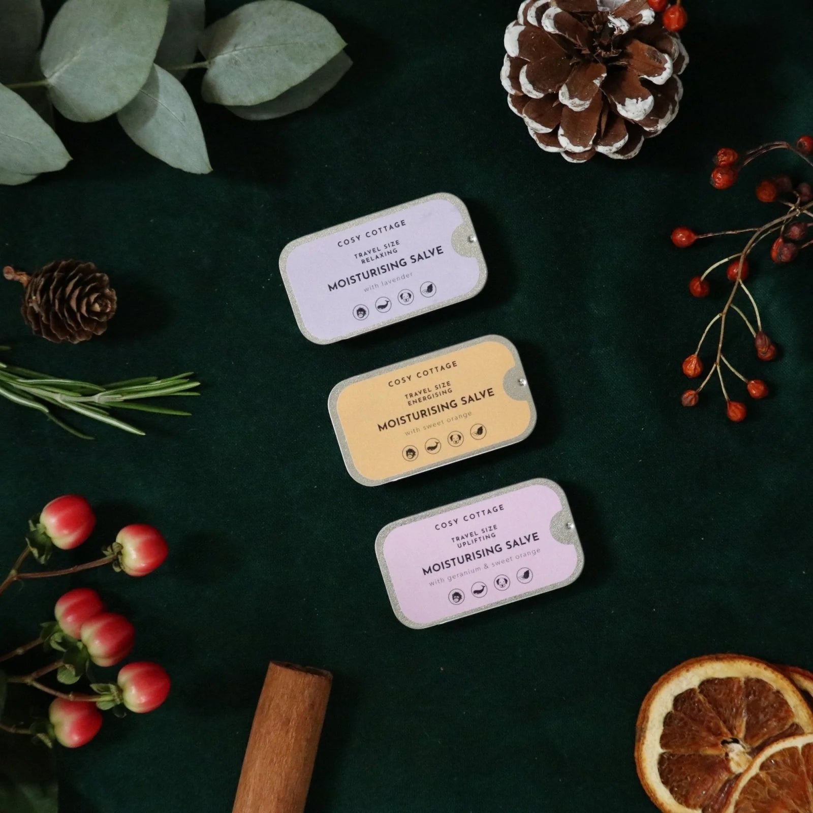 Three small containers of Cozy Cottages Moisturising Salve on a dark surface with decorative elements like pine cones and berries.