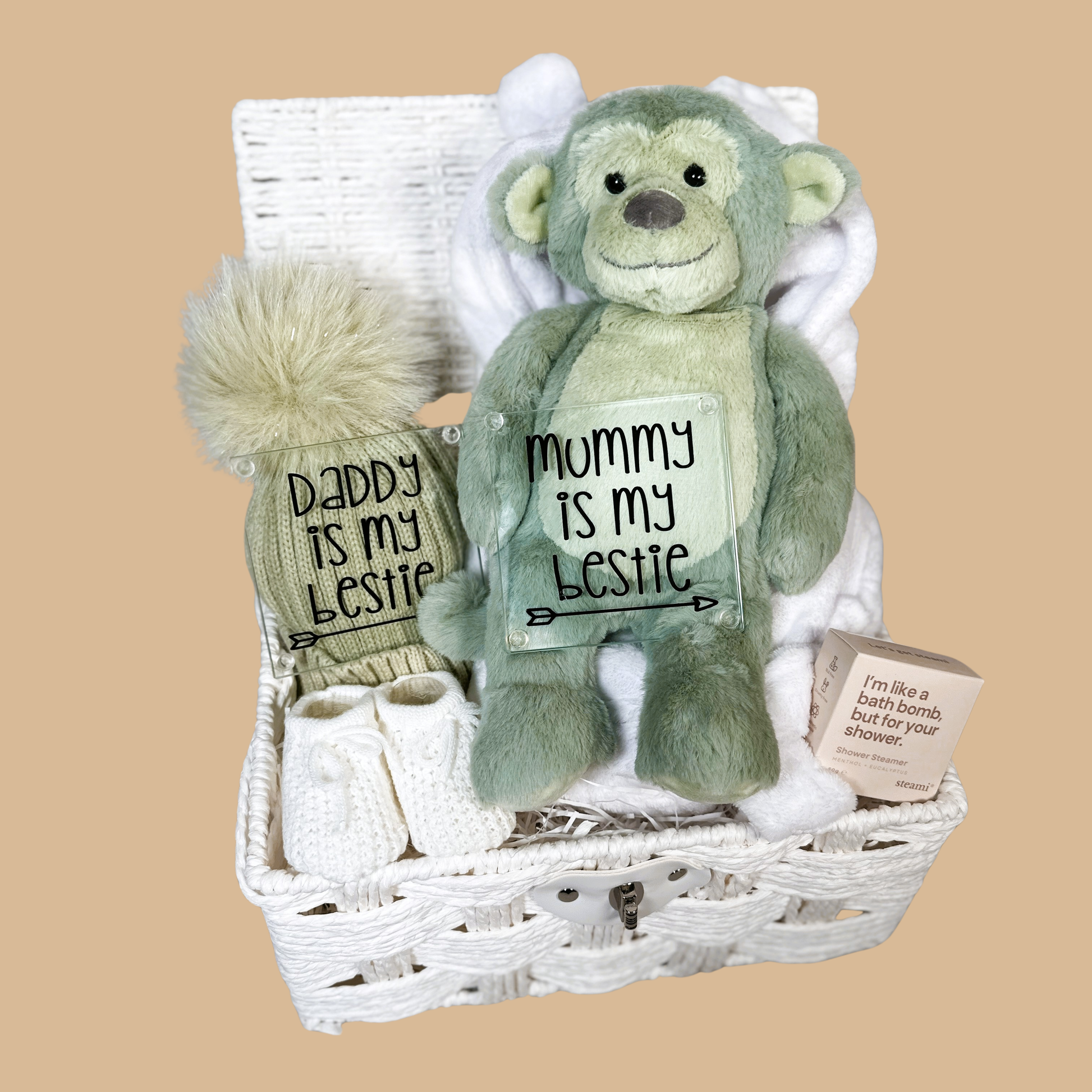 Neutral new baby hamper with a sage green plush monkey toy, a sage green baby pompom hat. Two new parents glass coasters, a pair of baby booties, shower steamer  in a white paper rope hamper.