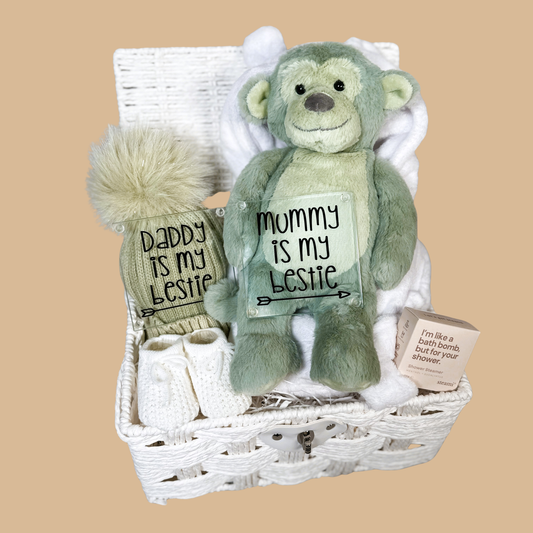 Neutral new baby hamper with a sage green plush monkey toy, a sage green baby pompom hat. Two new parents glass coasters, a pair of baby booties, shower steamer  in a white paper rope hamper.