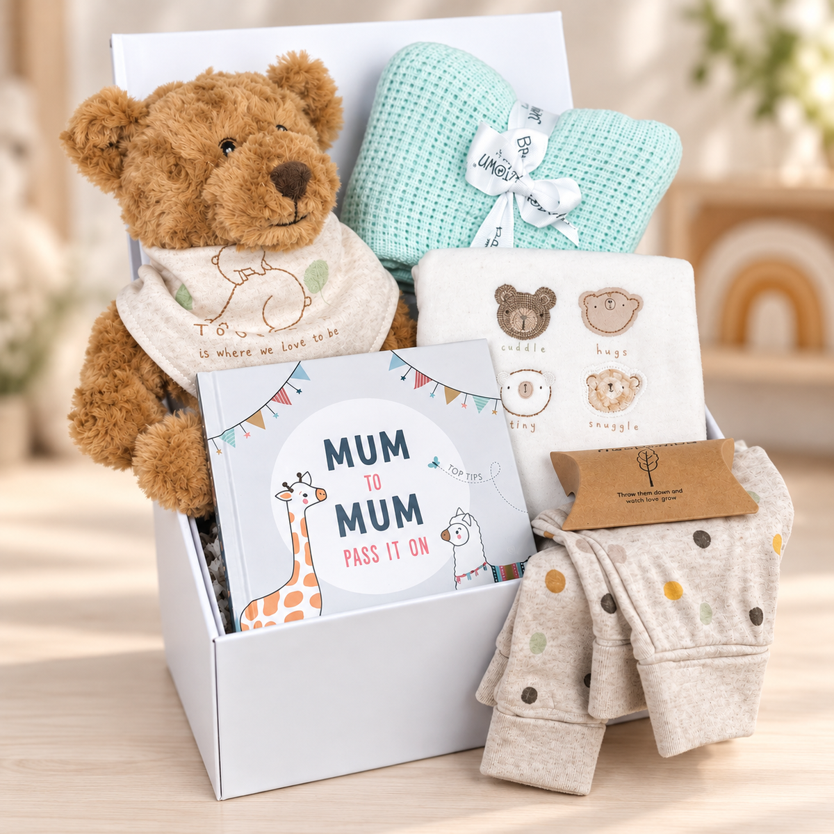A curated neutral new baby gift  arranged in a white baby keepsake box box on a light wood surface. The set includes a brown plush teddy bear wearing a cream bib, a mint green cellular baby blanket with a white ribbon, a white baby book titled 'Mum to Mum', a white baby bodysuit with embroidered animal faces, a pair of light beige baby baby trousers with multicolored dots, and a box o. Treebombs