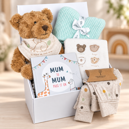 A curated neutral new baby gift  arranged in a white baby keepsake box box on a light wood surface. The set includes a brown plush teddy bear wearing a cream bib, a mint green cellular baby blanket with a white ribbon, a white baby book titled 'Mum to Mum', a white baby bodysuit with embroidered animal faces, a pair of light beige baby baby trousers with multicolored dots, and a box o. Treebombs