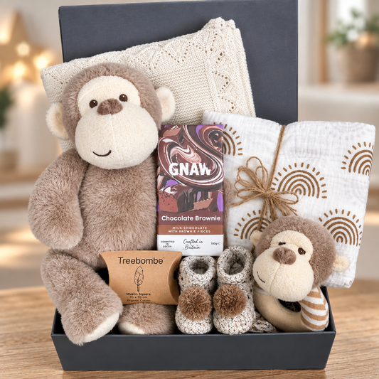 A baby gift hamper set in a baby keepsake box, featuring a Keeleco plush brown monkey toy and matching baby  ring rattle, a cream knit blanket, a white muslin cloth with brown sunburst patterns, a Treebomb package, crocheted baby booties and a GNAW chocolate brownie.
