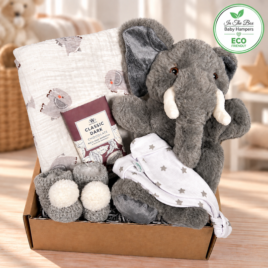 A baby hamper containing a grey elephant puppet made from recycled materials, a white baby knot hat with a grey stars print. A large white muslin square with an elephant print, a pair of crocheted baby bootees in grey with white pom poms and a bar of  chocolate.