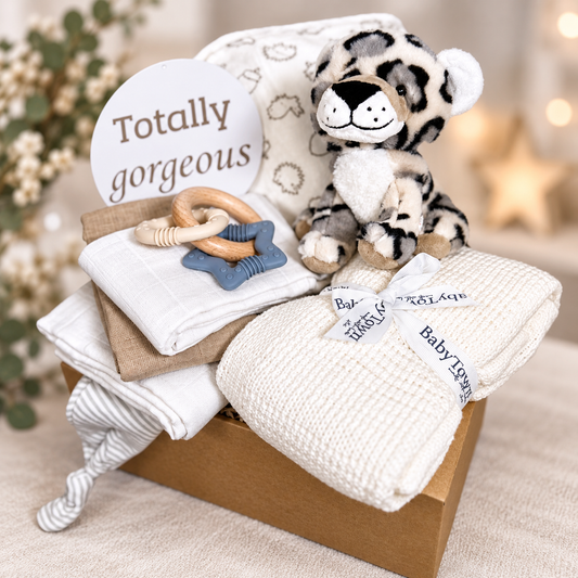 A curated neutral baby gift hamper set in a cardboard box featuring a plush tiger toy, a white cotton baby blanket, 3 baby muslin squares, a striped knotty baby hat, a wooden teething ring, a blue star-shaped teether, a cotton hooded baby towel and a round sign that reads 'Totally gorgeous'.