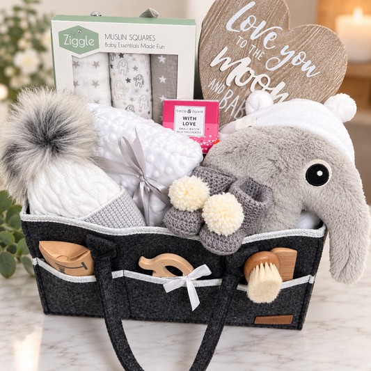 A curated baby gift hamper arranged in a dark grey felt nappy caddy on a light marble surface. The basket contains a plush grey elephant soft baby toy, a white baby pompom hat with  grey pom-poms, a white blanket tied with a grey ribbon, a box of Ziggle muslin squares, and wooden teething toys and bay hairbrush and comb set in the side pockets.