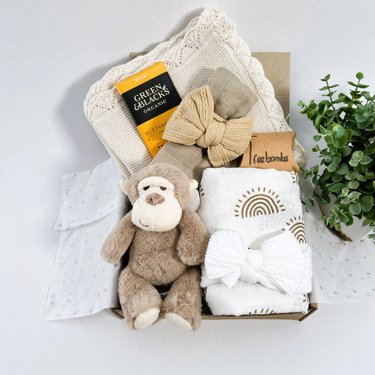 neutral new baby girl gift hamper containing a eura cotton baby blanket, a large white muslin with a brown sunshine print, two baby headbansa Keeleco Marley monkey 14cms soft baby toy and a bar of Green and Blacks organic butterscotch chocolate.