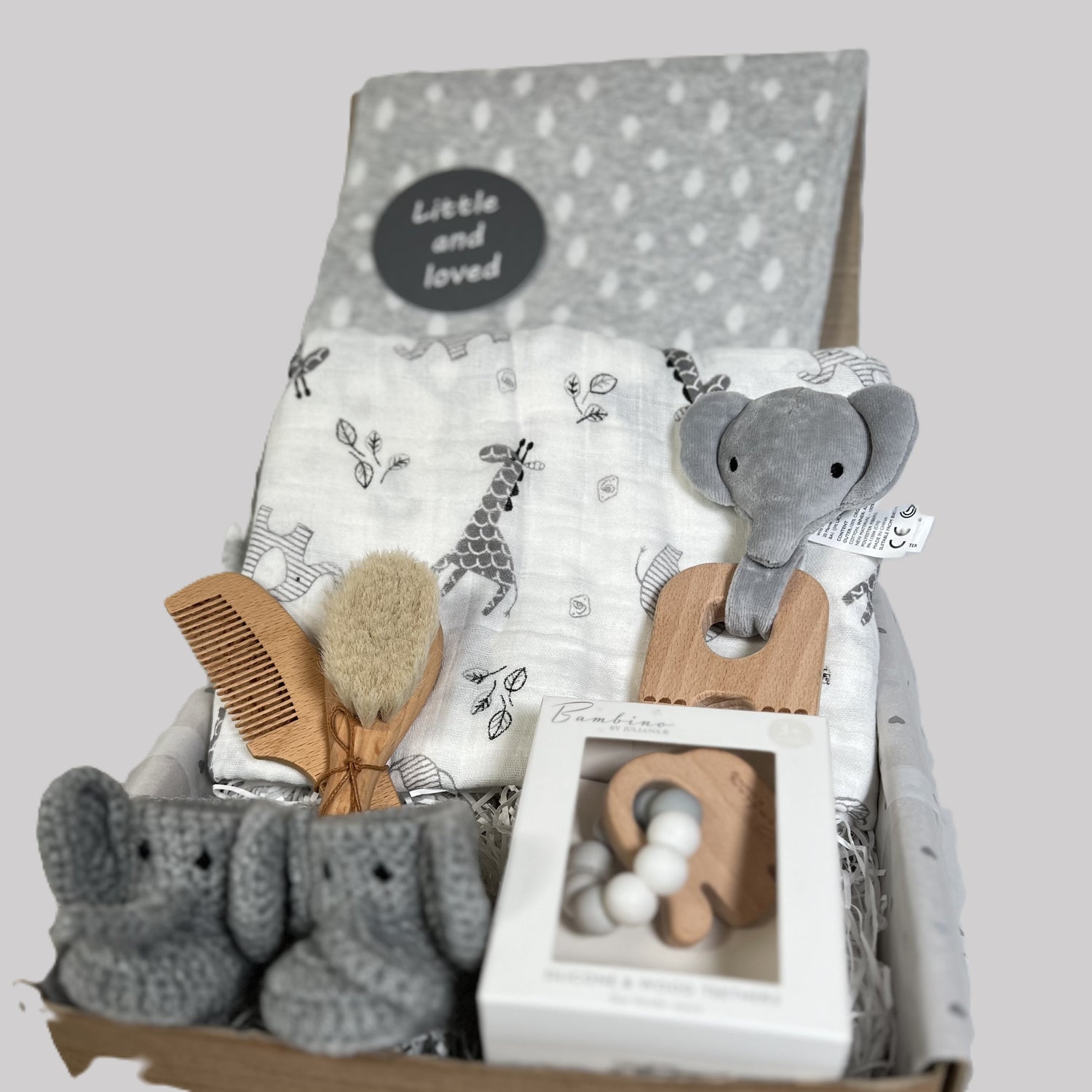 A stunning new baby hamper full of elephant baby essentials such aa a large white baby muslin with grey elephant and zebra print. Two elephant baby teething toys. A pair of crocheted baby booties with an elephant face, a grey organic cotton baby blanket and a baby hairbrush and comb set.