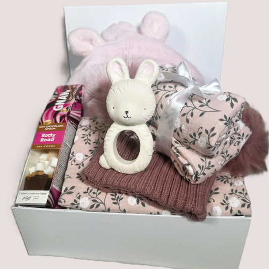 baby girl gift hamper with baby items including a bunny baby teether, swaddle blanket, muslins and chocolate stirrer on a light pink background