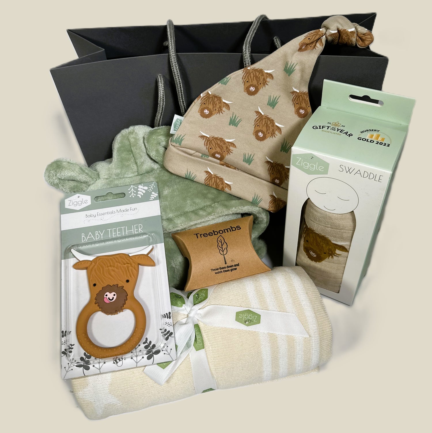 A new baby gift containing a cream and white cotton baby blanket , a highland cow print baby knot hat and baby swaddle. A highland cow silicone baby teether and a sage green baby dressing gown.