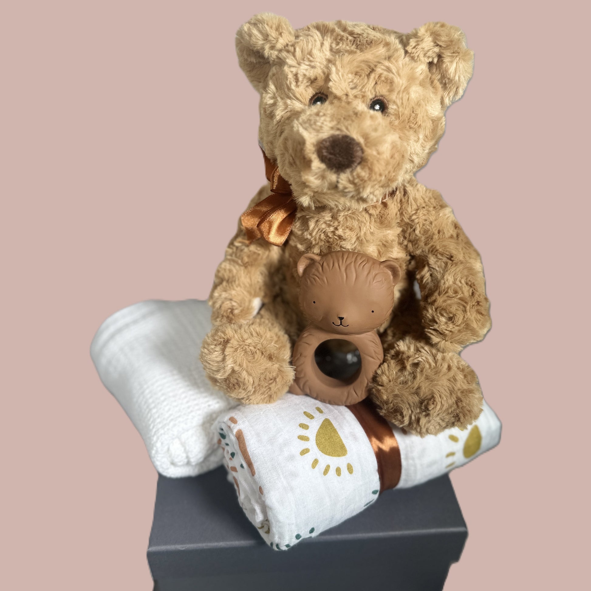 Two teddy bears, one larger and one smaller baby teether, sitting on a white blanket with a gold design against a beige background.