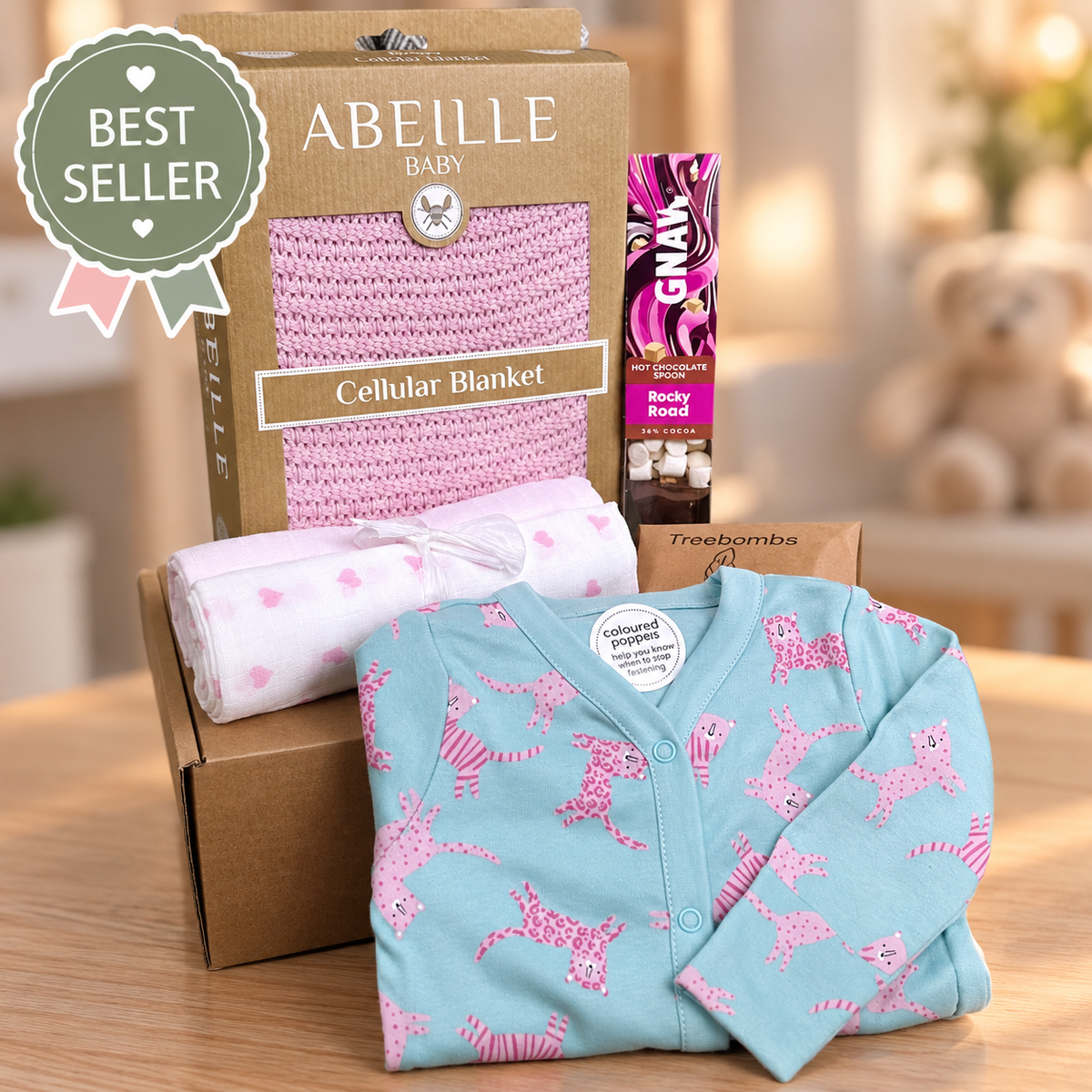 baby girl curated baby hamper gift with cotton cellular baby blanket in pink, a pink and blue cotton baby sleepsuit with pink leopard print, a Gnaw rocky road hot chocolate stirrer and two pink and white baby muslins.