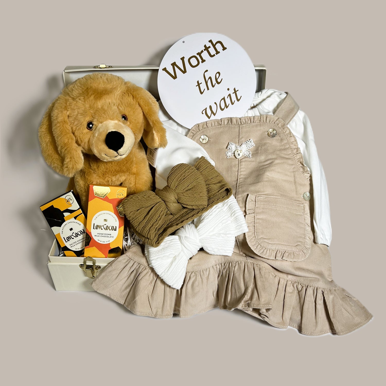 Baby Girl hamper in a cream case containing a neutral taupe pinafore dress and white frilled bodysuit, baby headbands, a soft labrador puppy soft toy and chocolate for mummy.