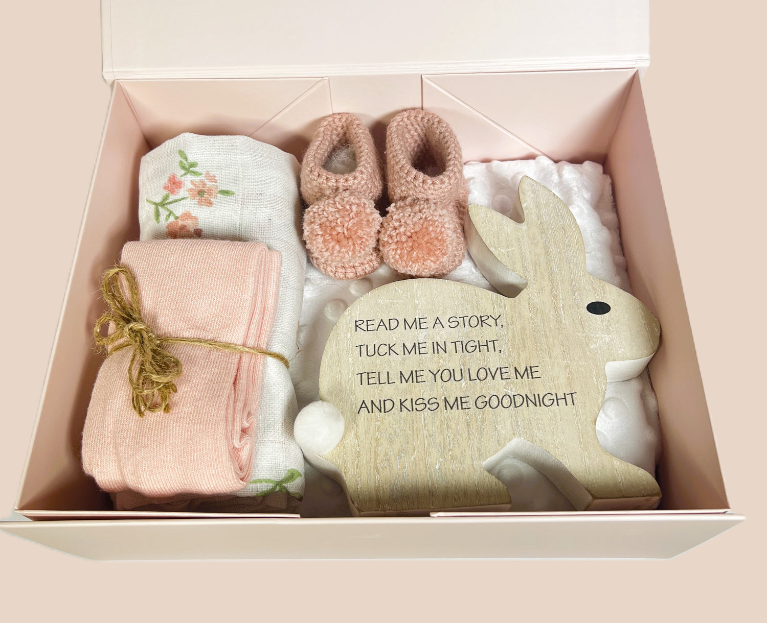 A new baby girl hamper with a corduroy pinafore dress and body suit with long sleeves and matching peach coloured baby tights. A pair of crocheted baby booties with pom poms. A white baby bubble blanket a, wood nursery ornament and a floral print baby muslin square.