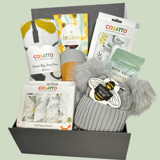Cosatto baby products including a hat, blanket, and bath soap in a box on a light green background