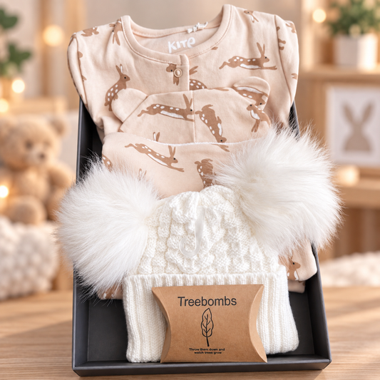 A curated baby hamper gift in a grey gift box, containing an organic cotton  baby romper, hat and bid with a hare print, a white knit beanie with fluffy white pom-poms, and a brown kraft gift box labelled Treebombs.