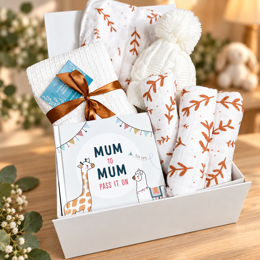 A baby gift hamper in a white baby keepsake box, featuring a white knit baby hat with a pom-pom, white muslins with orange leaf prints, a white waffle baby blanket cloth tied with a brown ribbon, and a book titled 'MUM TO MUM PASS IT ON'.