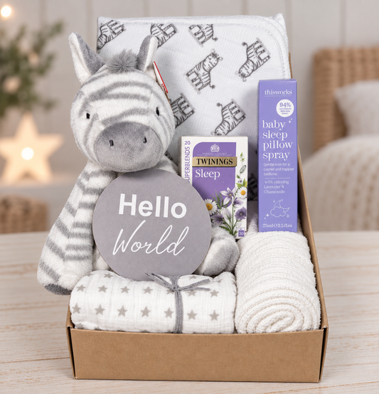 A  neutral baby gift hamper containing a plush zebra toy, a Twinings Sleep tea box, a bottle of This Works baby sleep pillow spray, a baby muslin, a folded white  and zebra print hooded baby towel, and a grey 'Hello World' sign, all arranged in a cardboard box on a wooden surface.