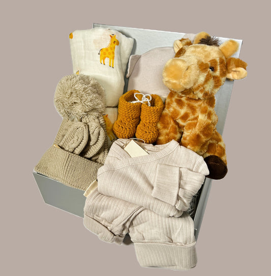 New baby hamper with an organic cotton baby outfit in taupe, a biscuits coloured baby pompom hat and matching mittens. A pair of mustard crocheted baby booties. A large giraffe print muslin  and Wilberry eco cuddlies giraffe soft baby toy.